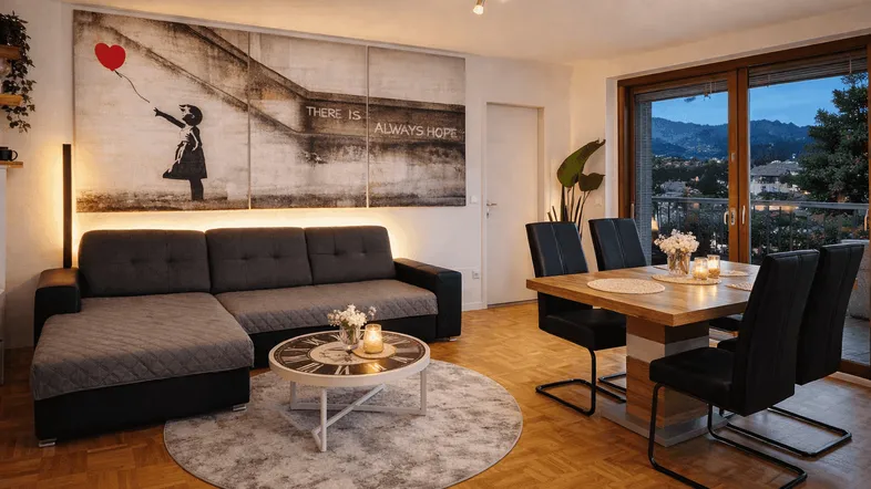 Gemütlicher Wohn- und Essbereich mit Banksy-Wandbild, Parkettboden und Zugang zum Balkon mit abendlicher Aussicht.