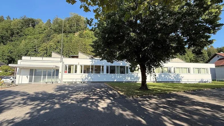 Modernes weißes Gebäude mit vielen Fenstern und großem Vorplatz, umgeben von grünen Hügeln.