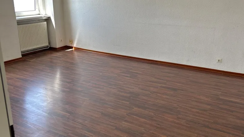 Helles, leeres Zimmer mit Laminatboden und einem Fenster, das viel Tageslicht hereinlässt.