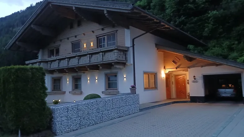 Traditionelles Alpenhaus bei Dämmerung mit beleuchtetem Eingangsbereich, Holzfassade und Gabionenmauer.