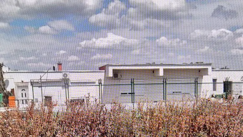 Gebäudeansicht von Weitem mit umliegenden Feldern unter bewölktem Himmel und einfacher Architektur.