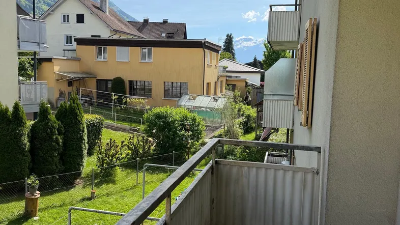 Blick vom Balkon auf einen grünen Garten, umliegende Häuser und die dahinterliegenden Berge.