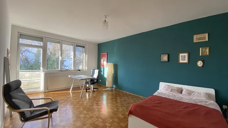 Helles Schlafzimmer mit Parkettboden, einer tealfarbenen Akzentwand und großem Fenster mit Grünblick.