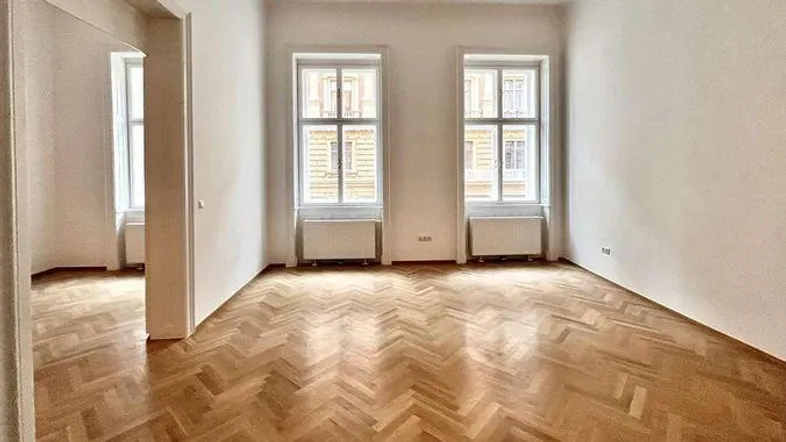 Geräumiges Zimmer mit klassischem Fischgrätparkett, weißen Wänden und zwei großen Fenstern für viel Tageslicht.