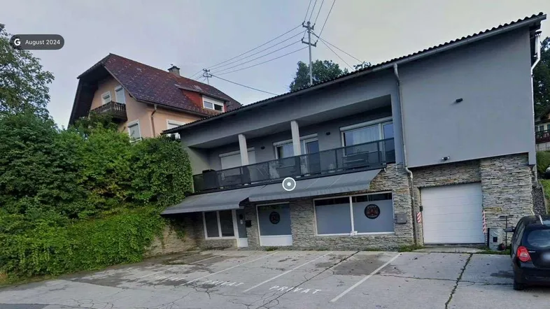 Graues Gebäude mit Steinverkleidung, Balkonen und Parkplätzen davor, in einer hügeligen Umgebung gelegen.