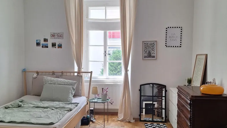 Helles Zimmer mit Holzboden, einem Bett, Sofa und einem Fenster mit Vorhängen.