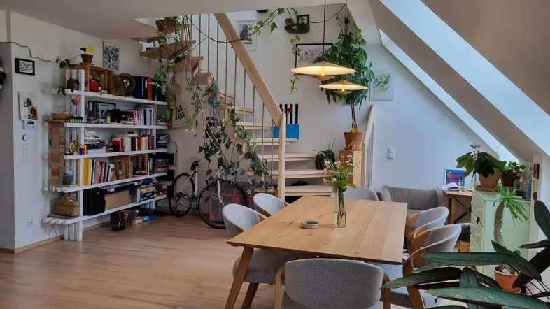 Geräumiger Wohn- und Essbereich mit Holztreppe, Bücherregalen und vielen Pflanzen unter einer Dachschräge.