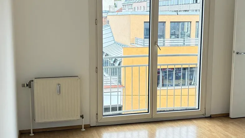 Helles Zimmer mit Parkettboden, Heizkörper und Zugang zu einem kleinen Balkon mit Stadtblick.