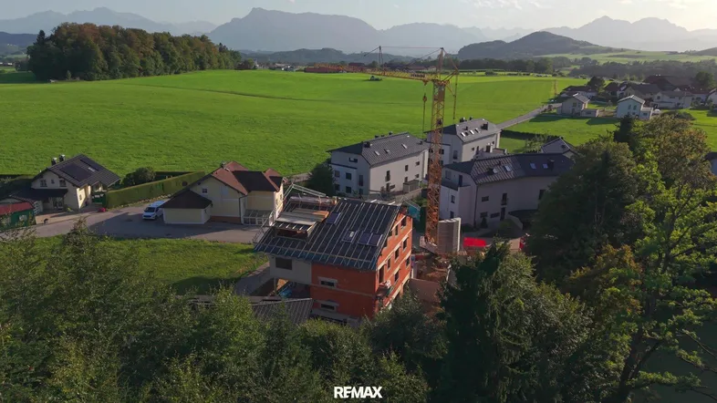 Luftaufnahme einer Baustelle mit einem im Bau befindlichen Haus, umgeben von grünen Feldern und Bergen.