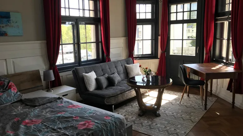 Geräumiges Wohnzimmer mit Parkettboden, bequemer Couch, Schreibtisch und großen Fenstern für viel Tageslicht.