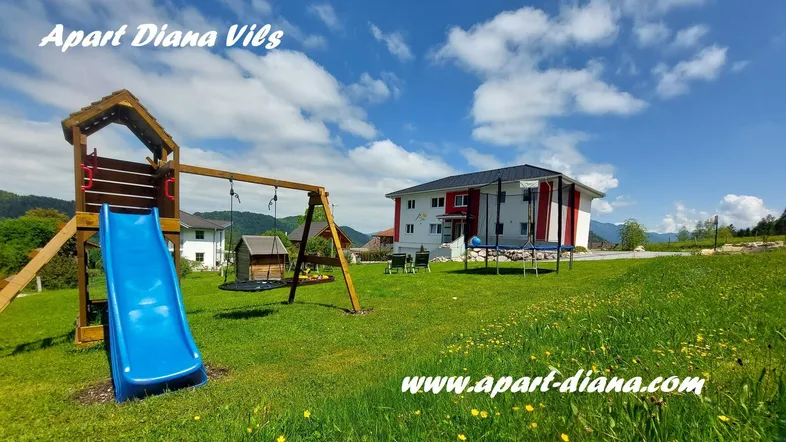 Großer Gartenbereich mit Spielplatz, Trampolin und Blick auf die Berge vor einem modernen Gebäude.