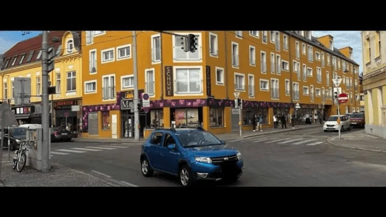 Gelbes Eckgebäude an einer belebten Straße mit Geschäften im Erdgeschoss und einem blauen Auto.