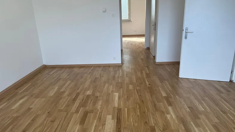 Geräumiger, leerer Raum mit hellem Holzparkett und weiß gestrichenen Wänden.