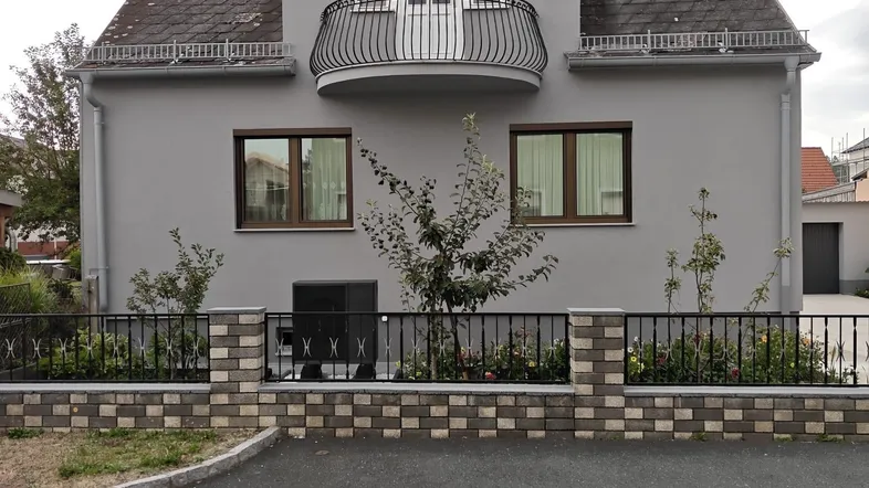 Graues Wohnhaus mit Mansarddach, kleinem Balkon und dekorativem Zaun an der Straße.