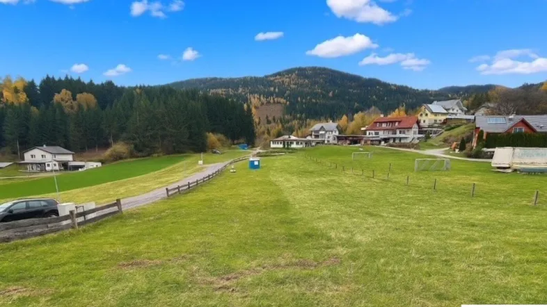Weitläufige grüne Wiese mit Blick auf umliegende Häuser und bewaldete Berge unter blauem Himmel.