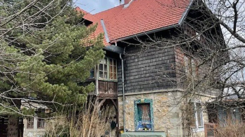Traditionelles Haus mit rotem Ziegeldach und Holzfassade, umgeben von Bäumen und Garten.