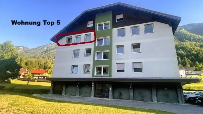 Mehrfamilienhaus mit markierter Wohnung Top 5 und Garagen im Erdgeschoss, umgeben von Bergen.