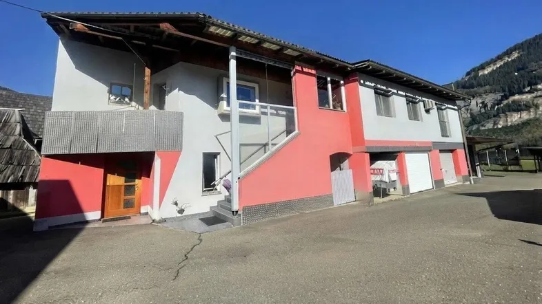 Gebäude mit roter und grauer Fassade, Balkon, Garagen und Carport, umgeben von ländlicher Landschaft mit Bergblick.
