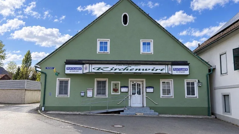 Grünes Gebäude mit Giebeldach und weißen Fenstern, das als Gasthof dient, unter blauem Himmel.