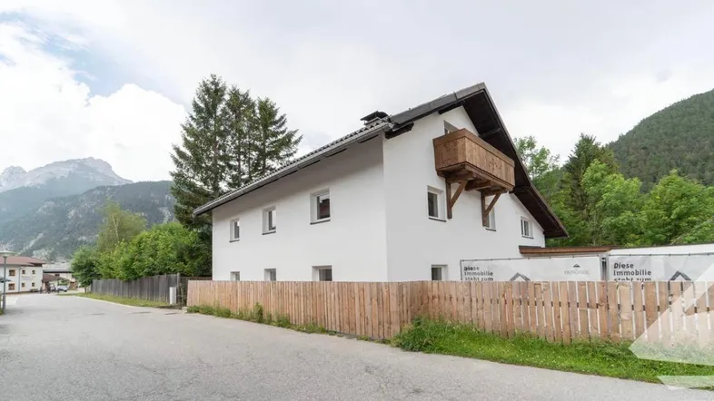 Weißes Haus mit kleinem Holzbalkon, umgeben von Bergen und Bäumen in ländlicher Umgebung.
