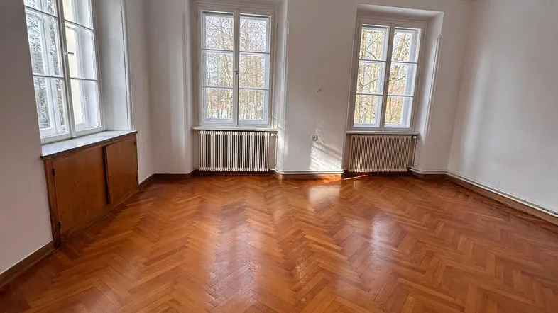 Geräumiges Zimmer mit Fischgrätparkett, weißen Wänden und großen Fenstern, die viel Tageslicht hereinlassen.