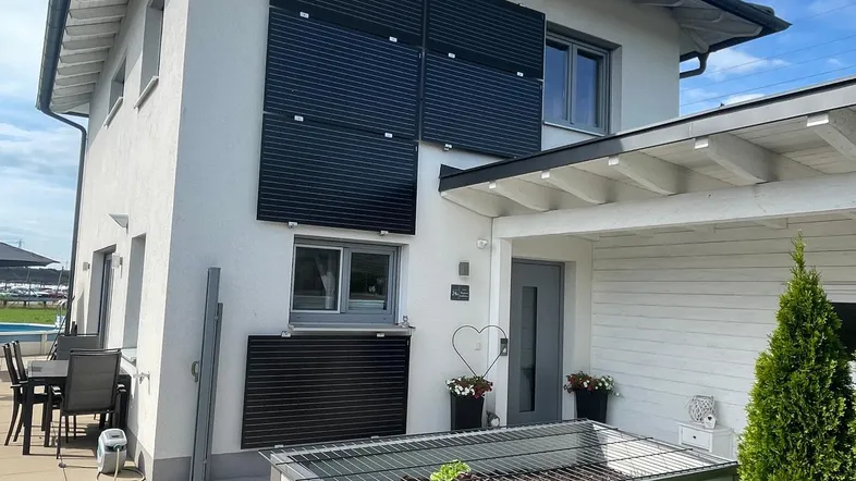 Moderne Hausfassade mit Terrasse, Grillbereich und Blick auf den Pool unter blauem Himmel.