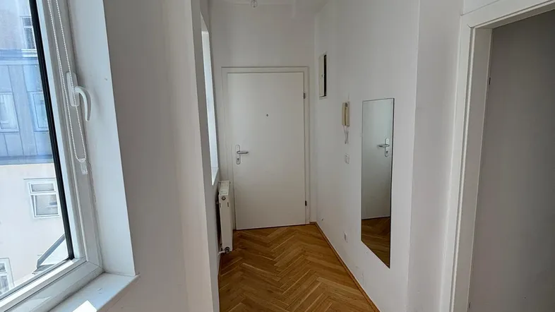 Heller Flur mit Fischgrätparkettboden und einem Fenster, das Tageslicht hereinlässt.