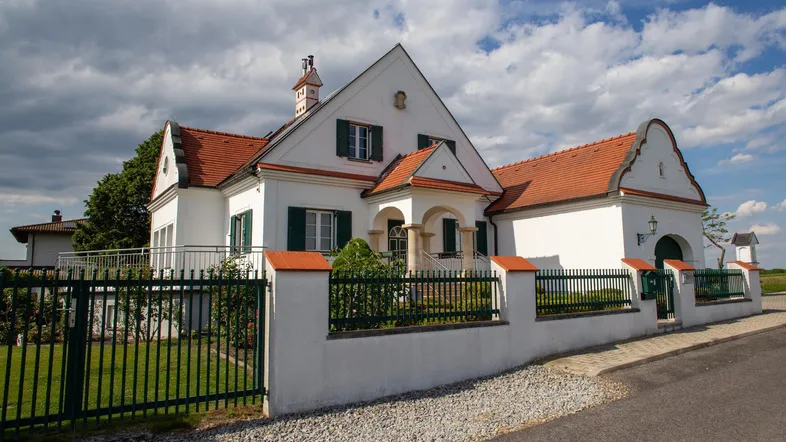 Weißes Haus mit rotem Ziegeldach, grünen Fensterläden und einem gepflegten Vorgarten mit Zaun.