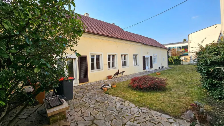 Hausfassade mit Steinterrasse und Rasenfläche im Gartenbereich unter blauem Himmel.