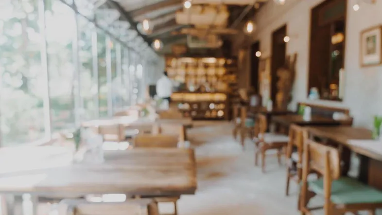 Heller Innenraum eines Restaurants mit Holztischen, Stühlen und großen Fensterfronten, die viel Tageslicht hereinlassen.