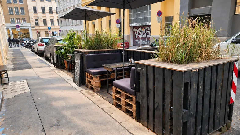Außenbereich eines Cafés mit Sitzgelegenheiten aus Palettenmöbeln, Sonnenschirmen und Pflanzen auf dem Bürgersteig.