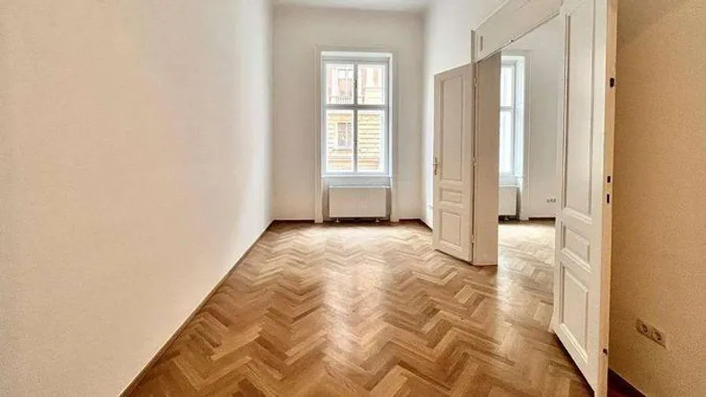 Leeres Zimmer mit Fischgrätparkett, weißen Wänden und einem Fenster, das viel Tageslicht hereinlässt.