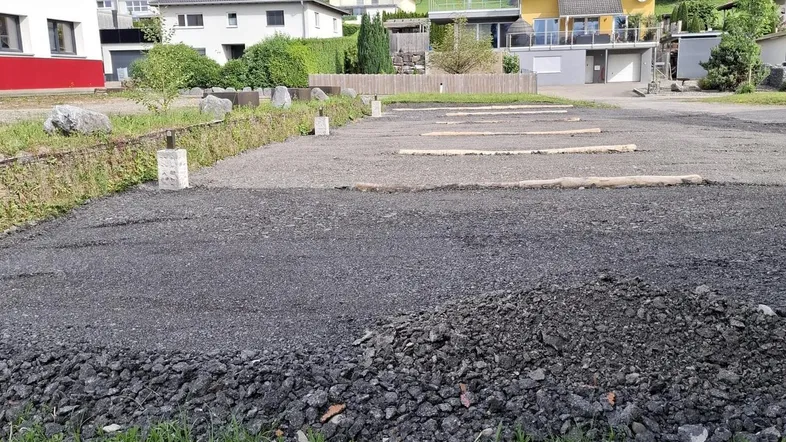 Ein Schotterparkplatz im Bau mit Blick auf bewaldete Hügel und umliegende Häuser in ländlicher Umgebung.