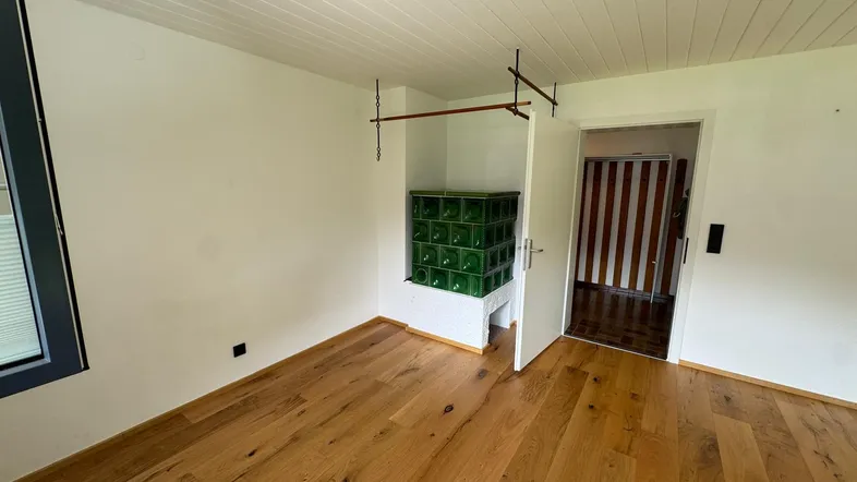 Helles Zimmer mit Holzboden und traditionellem grünem Kachelofen, der Wärme spendet.