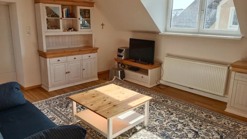 Gemütliches Wohnzimmer mit Dachschräge, Teppich und traditionellen Holzmöbeln, hell durch das Fenster.
