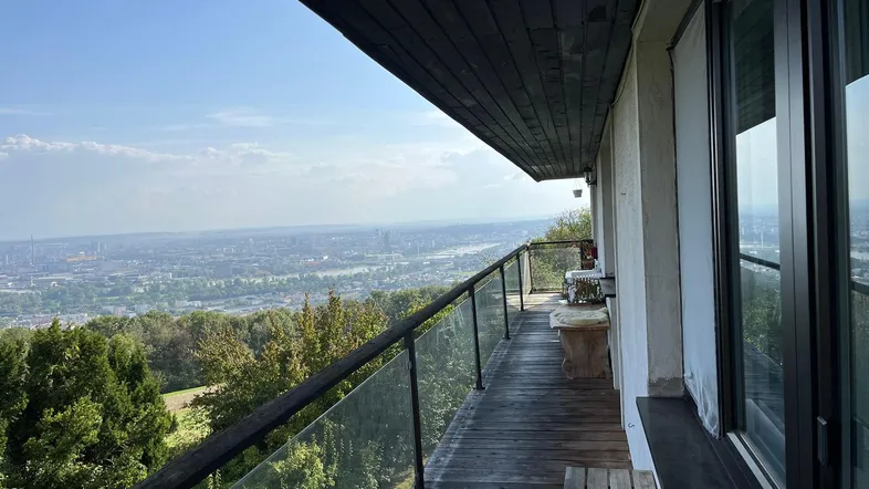 Langer Balkon mit Holzfußboden und Glasgeländer, der einen weiten Blick über die Stadt und Landschaft bietet.