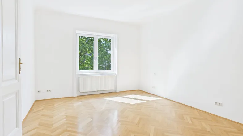 Helles, leeres Zimmer mit Fischgrätparkett und Blick ins Grüne durch das Fenster.