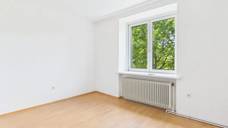 Helles, leeres Zimmer mit Parkettboden und Fenster mit Blick ins Grüne.