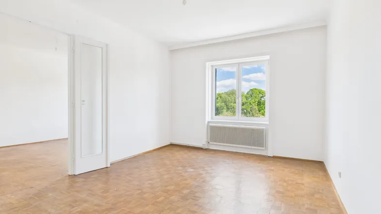 Helles Zimmer mit Parkettboden, weißen Wänden und Blick ins Grüne durch das Fenster.