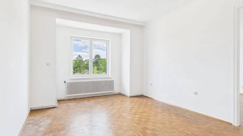 Helles Zimmer mit Parkettboden, weißen Wänden und Blick ins Grüne durch das Fenster.