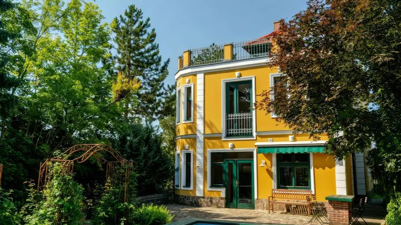 Gelbe Villa mit Pool im Garten und üppiger Vegetation unter blauem Himmel.
