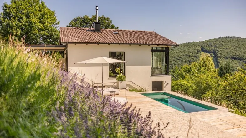 Modernes Haus mit Pool und Holzdeck, umgeben von grüner Natur und Hügellandschaft.