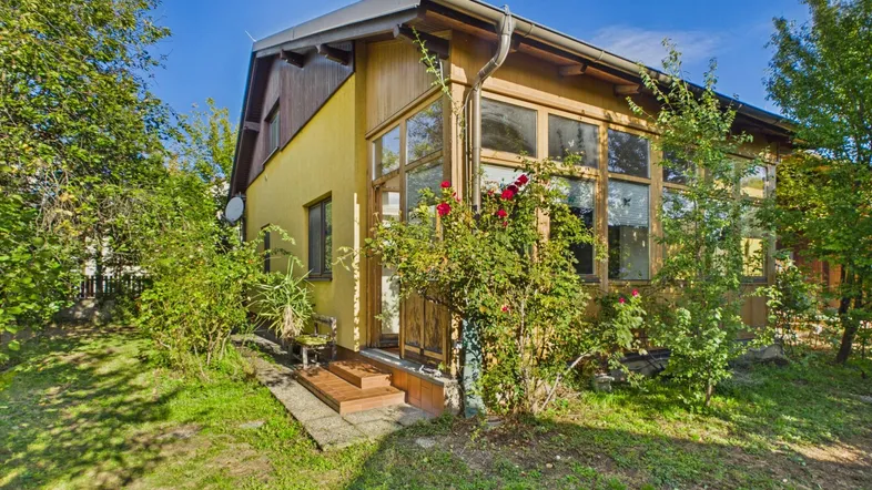 Gelbes Haus mit verglastem Anbau und umgebendem Garten unter blauem Himmel.