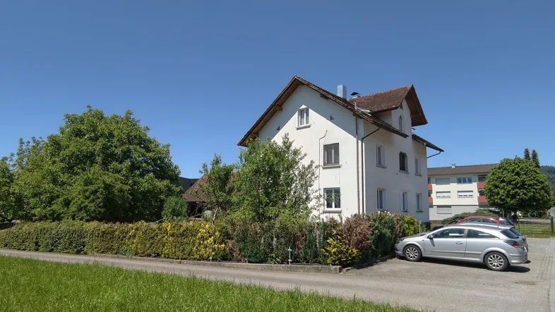Weißes Mehrfamilienhaus mit Satteldach und umliegender Grünfläche unter blauem Himmel.