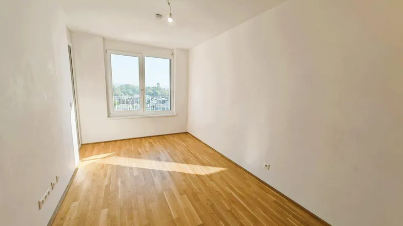 Helles Zimmer mit Parkettboden und großen Fenstern, das viel Tageslicht hereinlässt.