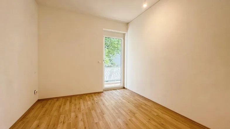 Helles, leeres Zimmer mit Holzboden und Zugang zu einem Balkon, ideal als Schlafzimmer.