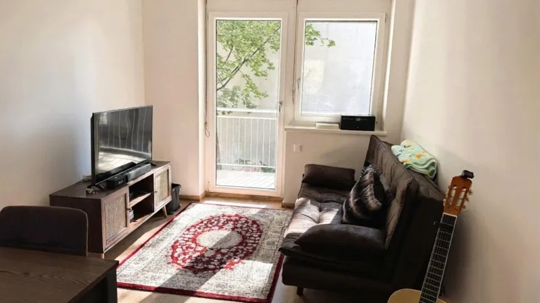 Helles Wohnzimmer mit Sofa, Fernseher und Zugang zu einem kleinen Balkon.