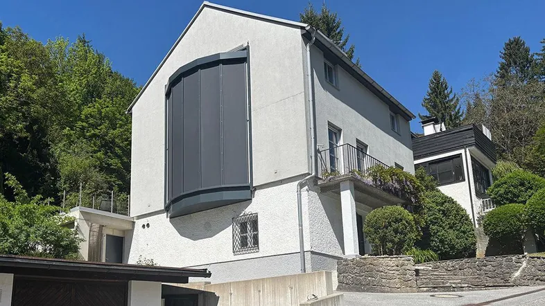 Modernes Haus mit markanter, geschwungener Fensterfront und Rollläden, umgeben von grünen Hügeln unter blauem Himmel.