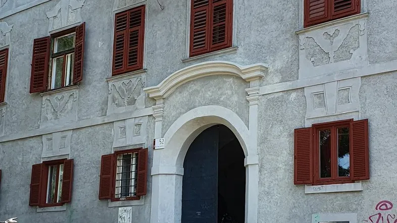 Historisches Gebäude mit grauer Fassade, roten Fensterläden und einem verglasten Balkon.