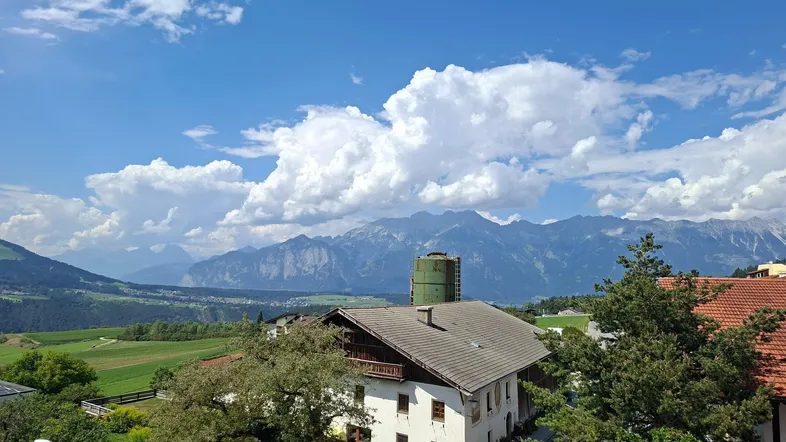 Traditionelles Haus mit Bergblick, umgeben von grünen Feldern und Bäumen unter blauem Himmel.